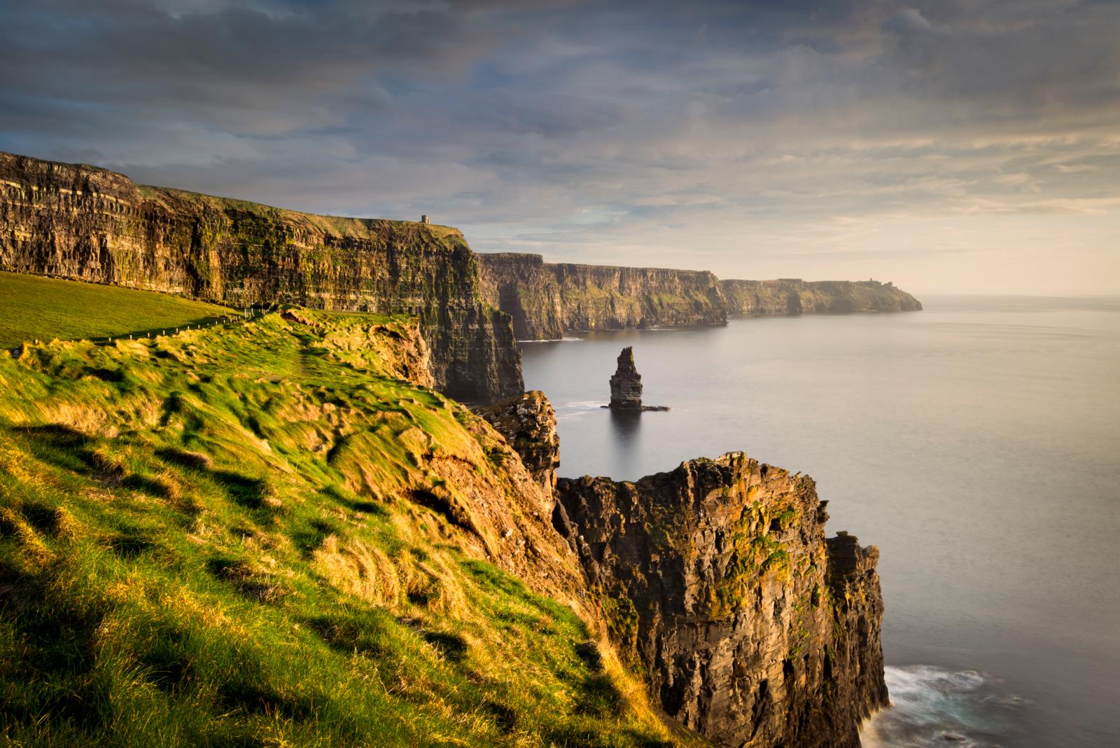 Een weids panorama bij golden hour van de Cliffs of Moher in Ierland, gezien vanaf een grasgroene landtong. De iconische Branaunmore zeestapel staat in de kalme zee, met O'Brien's Tower in de verte onder een dramatische, deels bewolkte hemel.