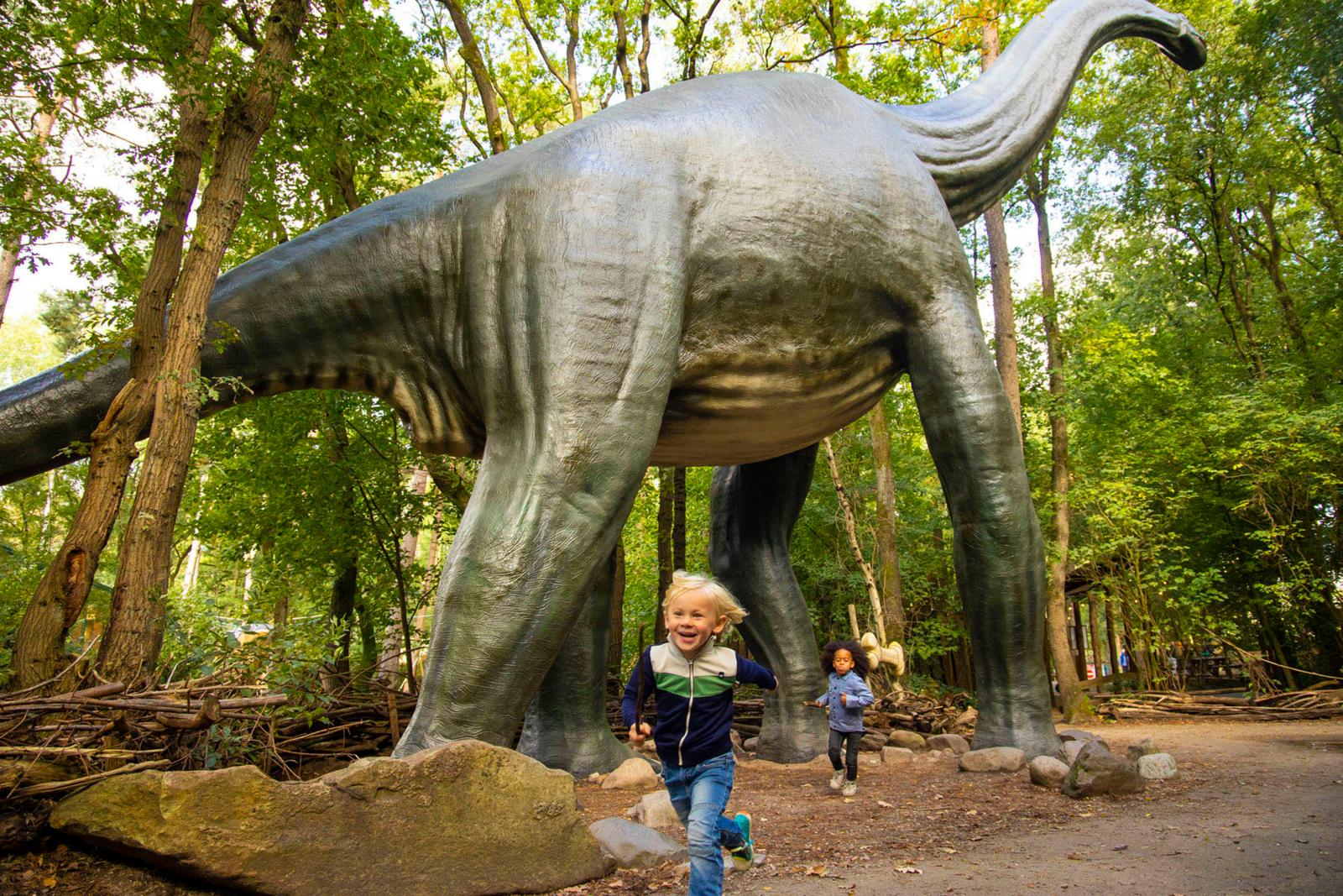 Twee lachende kinderen rennen door het bos in het Dinobos van DierenPark Amersfoort, met op de achtergrond een enorme, levensgrote replica van een Brachiosaurus dino.
