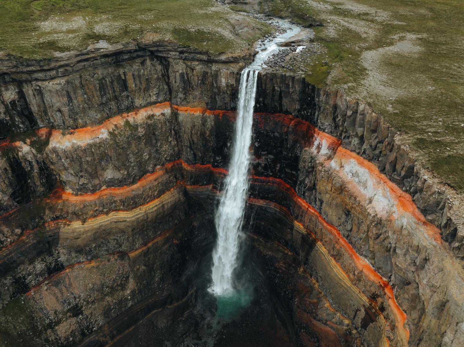 Bovenaanzicht van de Hengifoss waterval die langs een steile rotswand met opvallende horizontale rode en zwarte basaltlagen naar beneden klettert.
