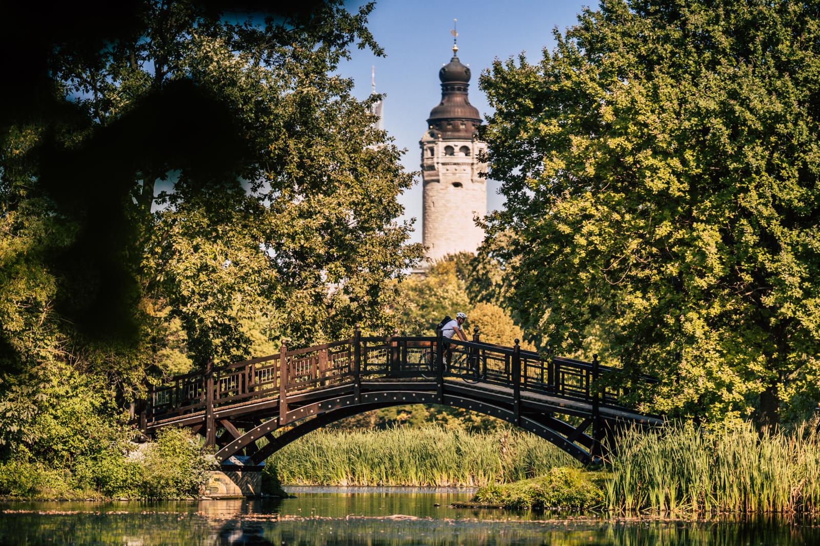 Een houten brug over het water in het groene Johannapark met de toren van het Neues Rathaus in de verte.