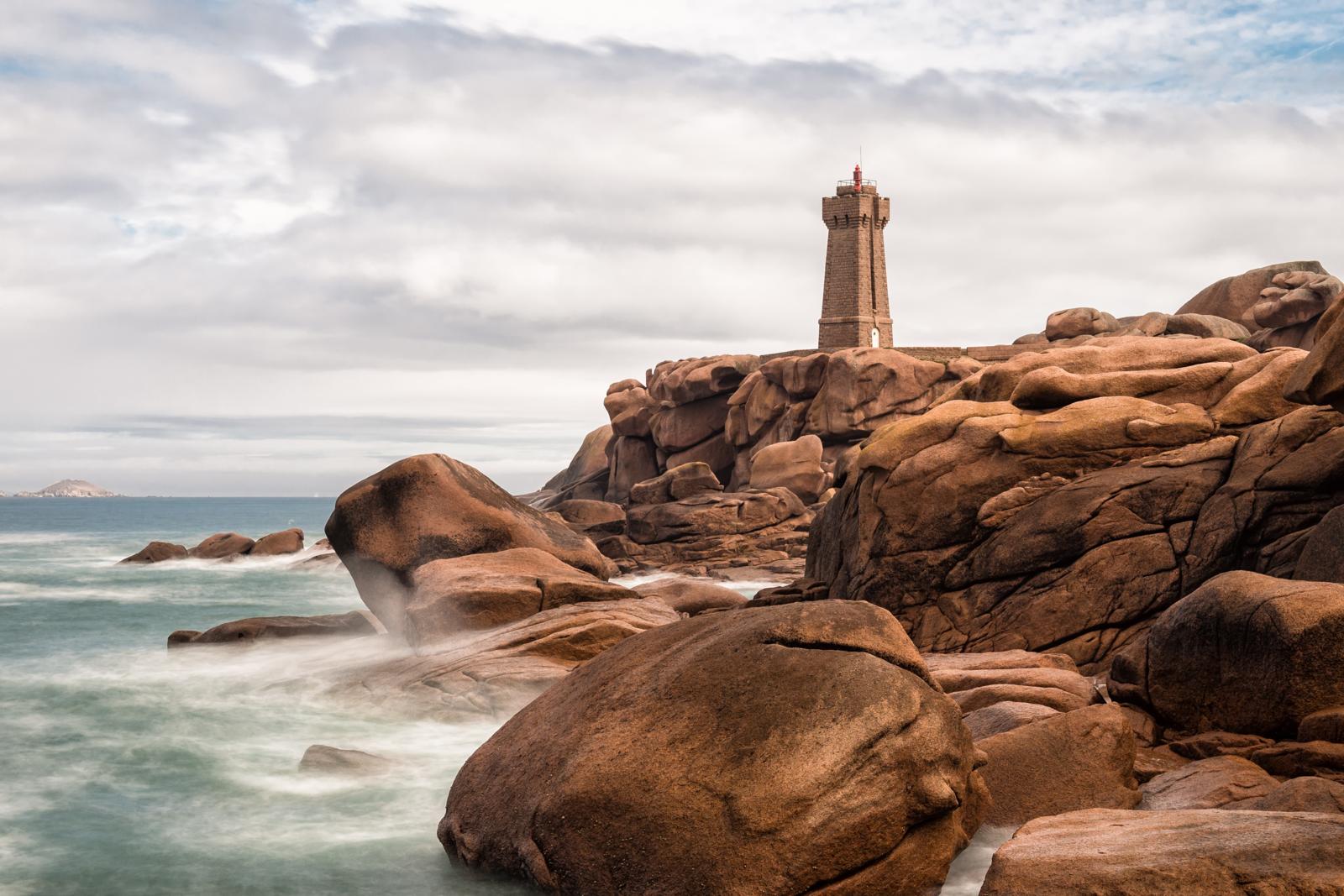 Een lange-sluitertijd foto toont de stenen Phare de Saint-Guirec (Ploumanac'h) op een klif van grote, gladde roze granieten rotsblokken, met een zijdeachtige zee die ertegenaan kabbelt onder een bewolkte hemel.