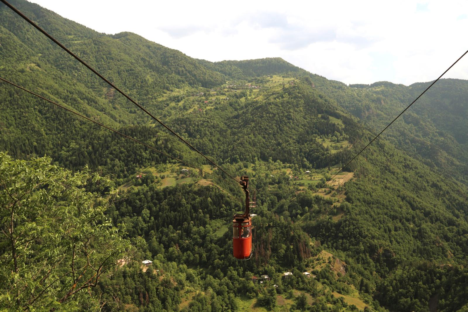 Een kleine, rode retro-kabelbaan hangt aan dikke kabels boven een uitgestrekt, dichtbebost berglandschap met verspreide huisjes in Georgië.