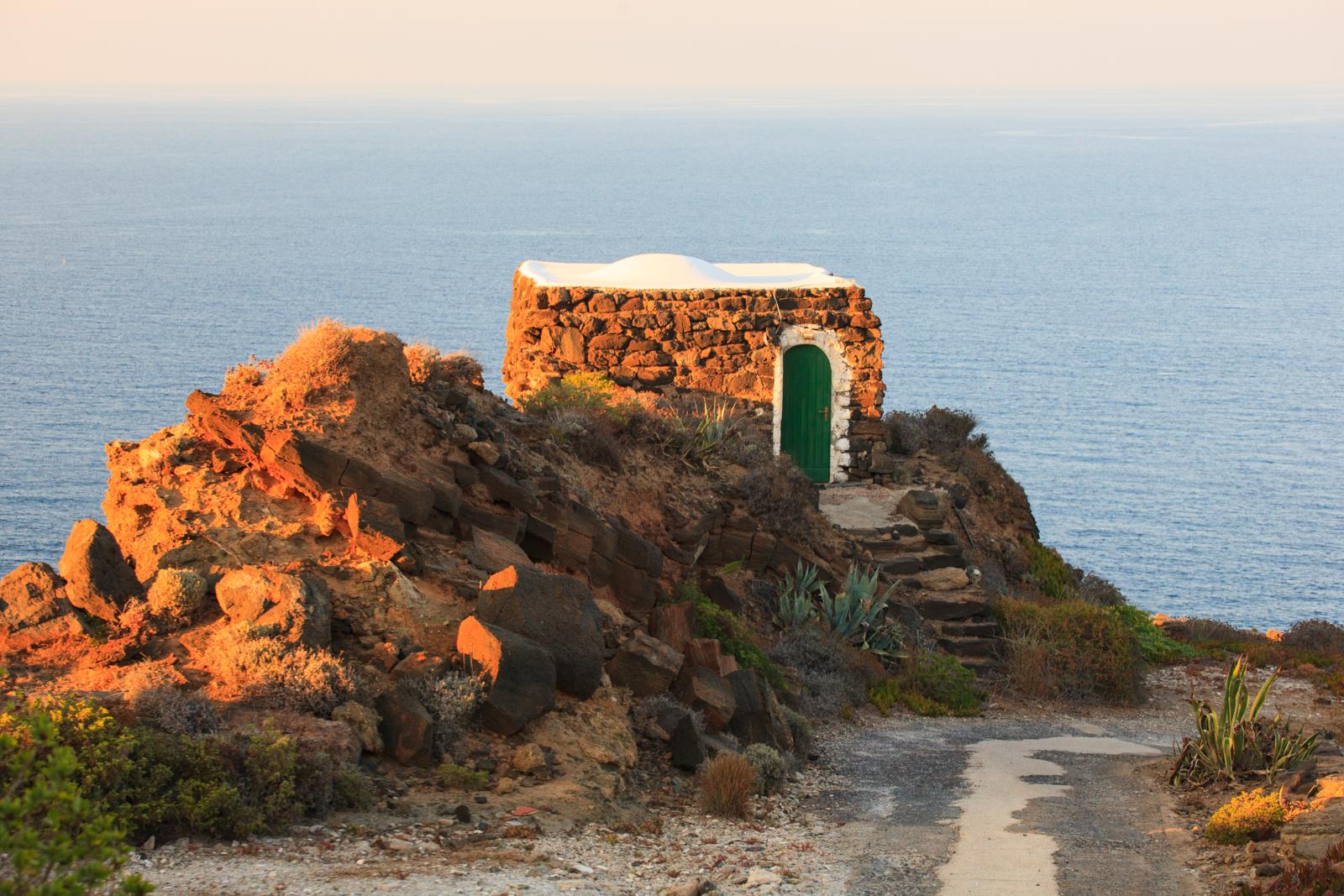 Een klein huisje gebouwd van donkere vulkanische steen met een wit koepeldak en een groene deur, gelegen op een klif aan de kust van Pantelleria bij zonsondergang.