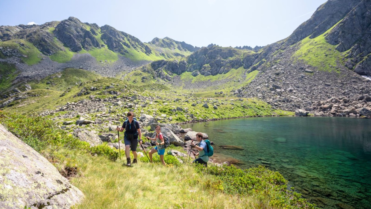 Drie bergwandelaars met wandelstokken passeren een turquoise bergmeer in het ruige landschap van Oostenrijk.