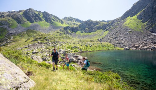 Drie bergwandelaars met wandelstokken passeren een turquoise bergmeer in het ruige landschap van Oostenrijk.