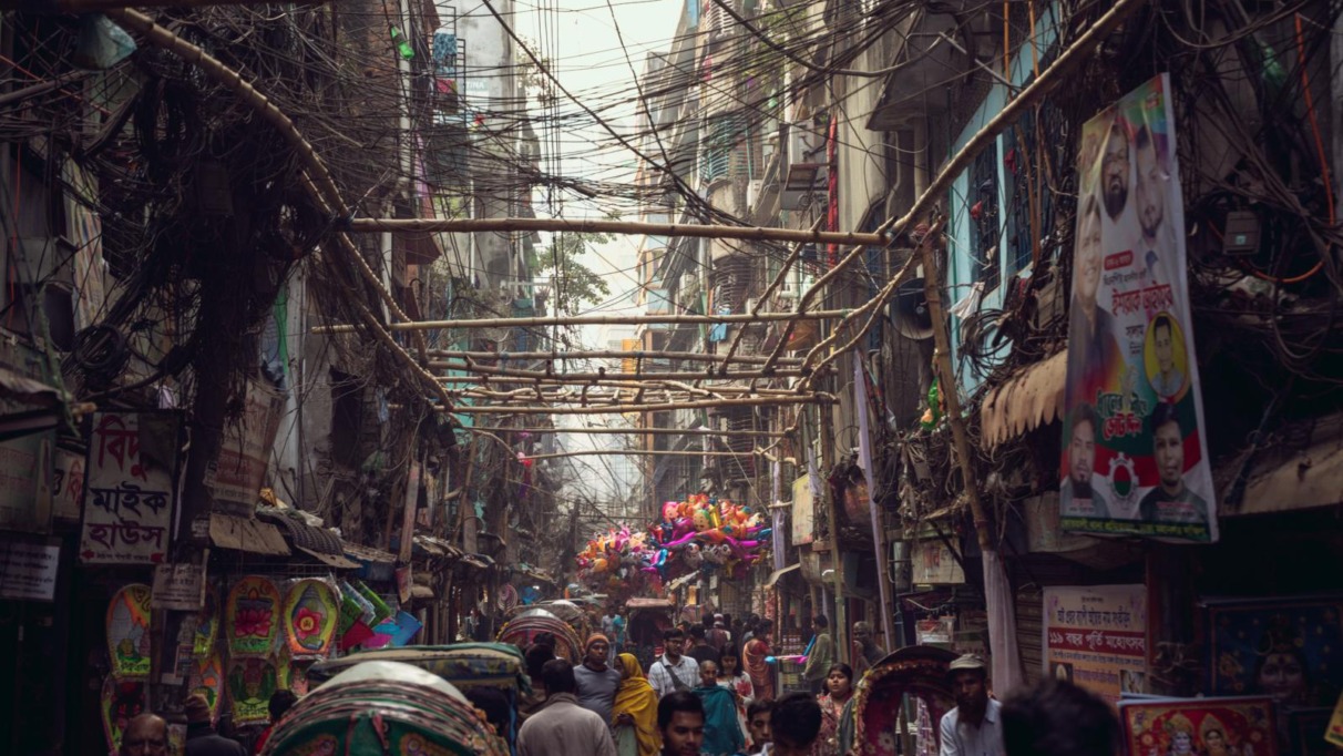 In Shankhari Bazaar, de eeuwenoude hindoestraat van oud-Dhaka, schuifelen riksja’s, marktkooplui en voetgangers door een wirwar van kabels, nauwe steegjes en dicht opeengepakte huizen die het chaotische ritme van Bangladesh’ hoofdstad voelbaar maken.