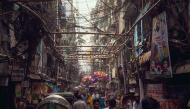 In Shankhari Bazaar, de eeuwenoude hindoestraat van oud-Dhaka, schuifelen riksja’s, marktkooplui en voetgangers door een wirwar van kabels, nauwe steegjes en dicht opeengepakte huizen die het chaotische ritme van Bangladesh’ hoofdstad voelbaar maken.