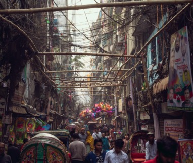 In Shankhari Bazaar, de eeuwenoude hindoestraat van oud-Dhaka, schuifelen riksja’s, marktkooplui en voetgangers door een wirwar van kabels, nauwe steegjes en dicht opeengepakte huizen die het chaotische ritme van Bangladesh’ hoofdstad voelbaar maken.