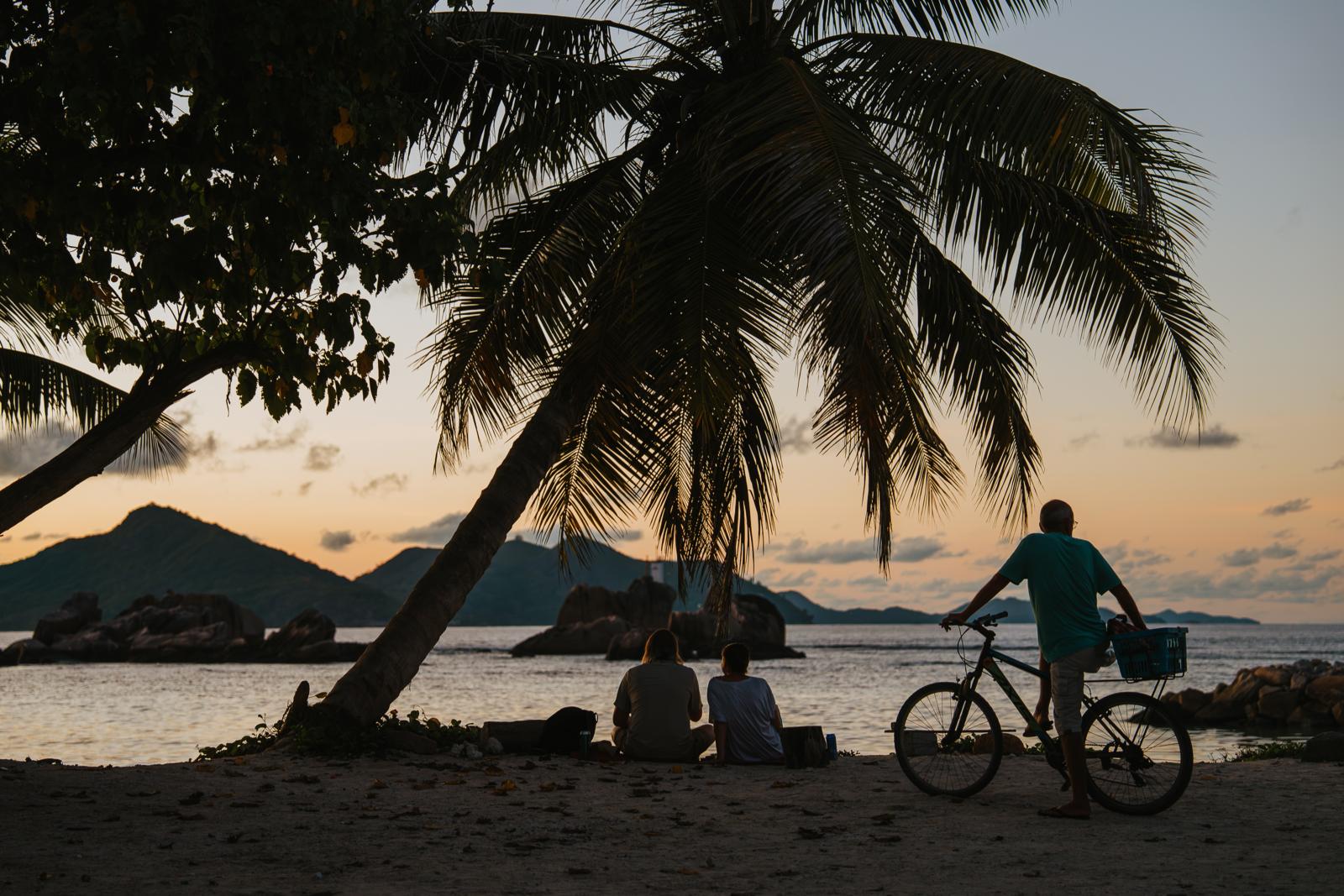 Silhouetten van reizigers en een fietser onder palmbomen aan het strand van La Digue tijdens zonsondergang