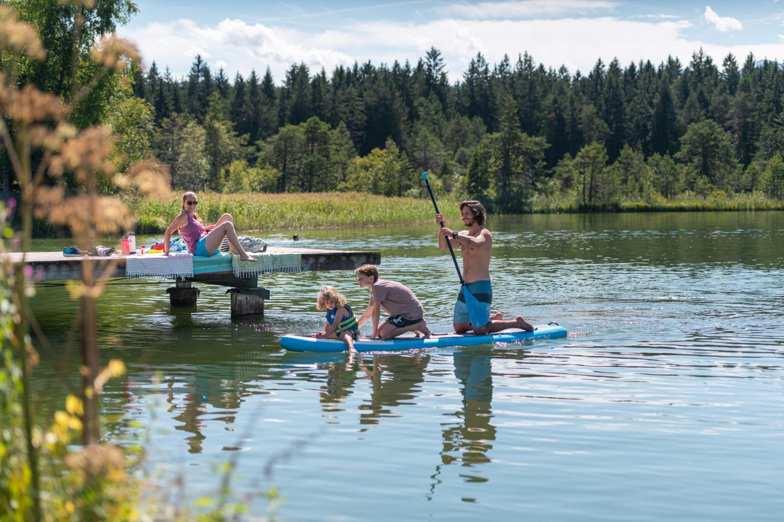 Een gezin op een SUP-board op de Wildsee in Seefeld, met een toekijkende vrouw op een houten steiger.