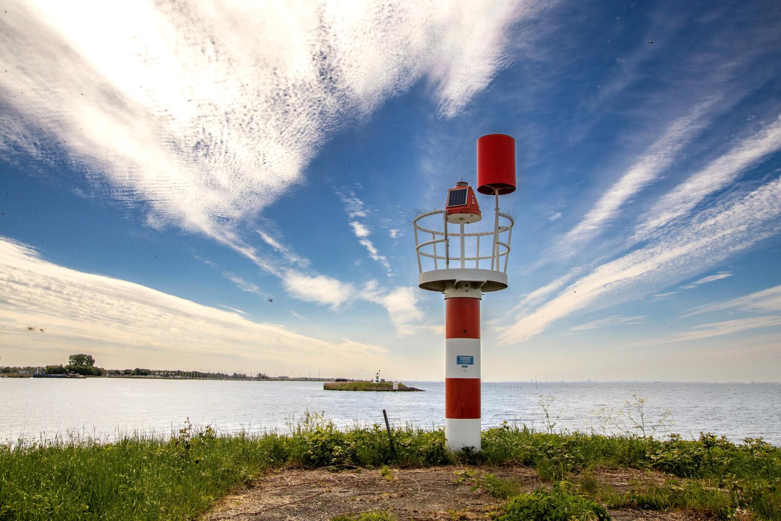 Een rood-wit gestreept baken aan de waterkant onder een spectaculaire blauwe lucht met vederwolken