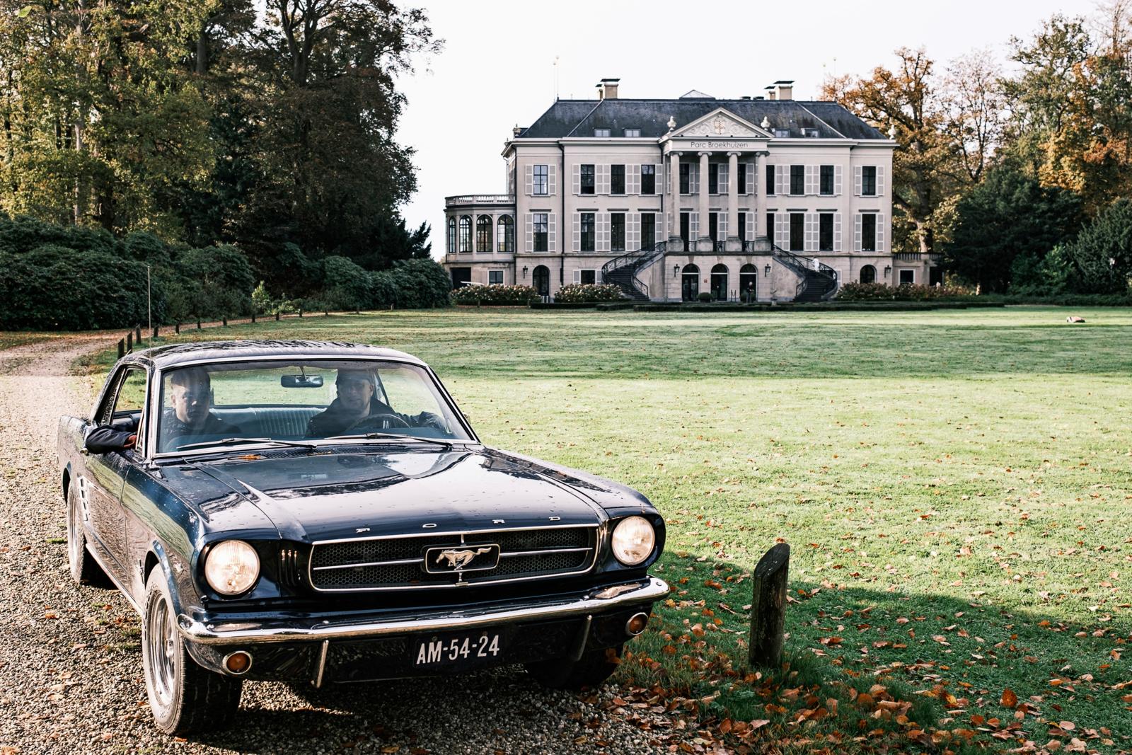 Een klassieke donkerblauwe Ford Mustang op een grindpad voor het imposante witte landhuis van Parc Broekhuizen in Leersum.