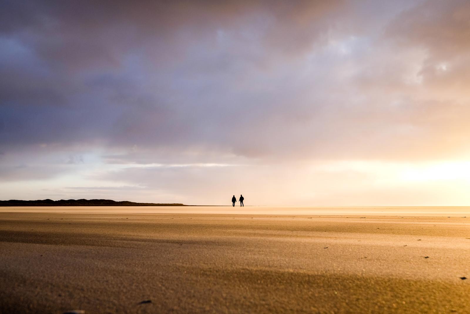 Twee wadlopers wandelen als silhouet over een goudkleurige zandplaat richting de horizon bij het krieken van de dag.