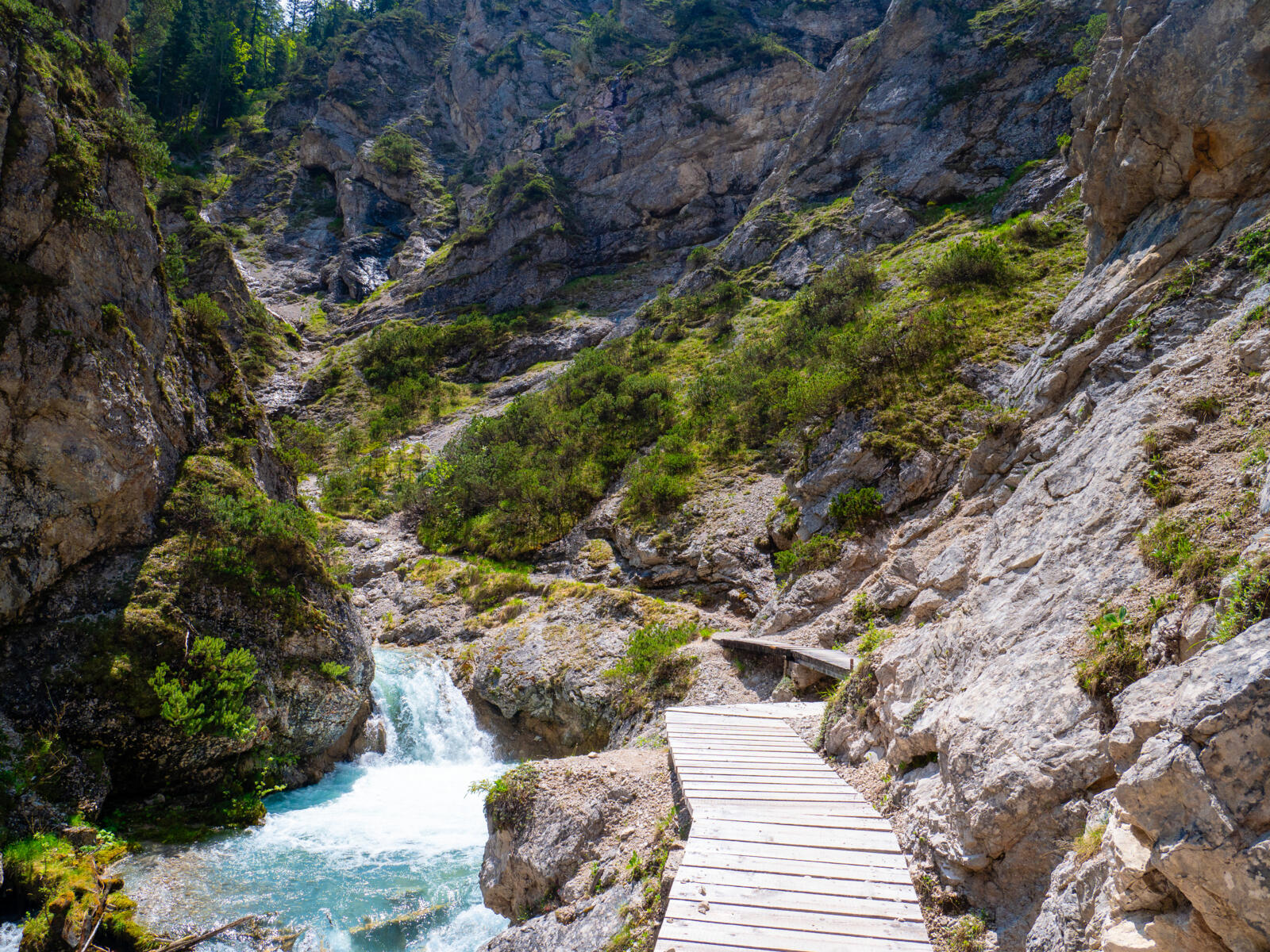 Een houten wandelpad loopt door een rotsachtige kloof langs een stromende waterval en een turquoise bergbeek in de Alpen.