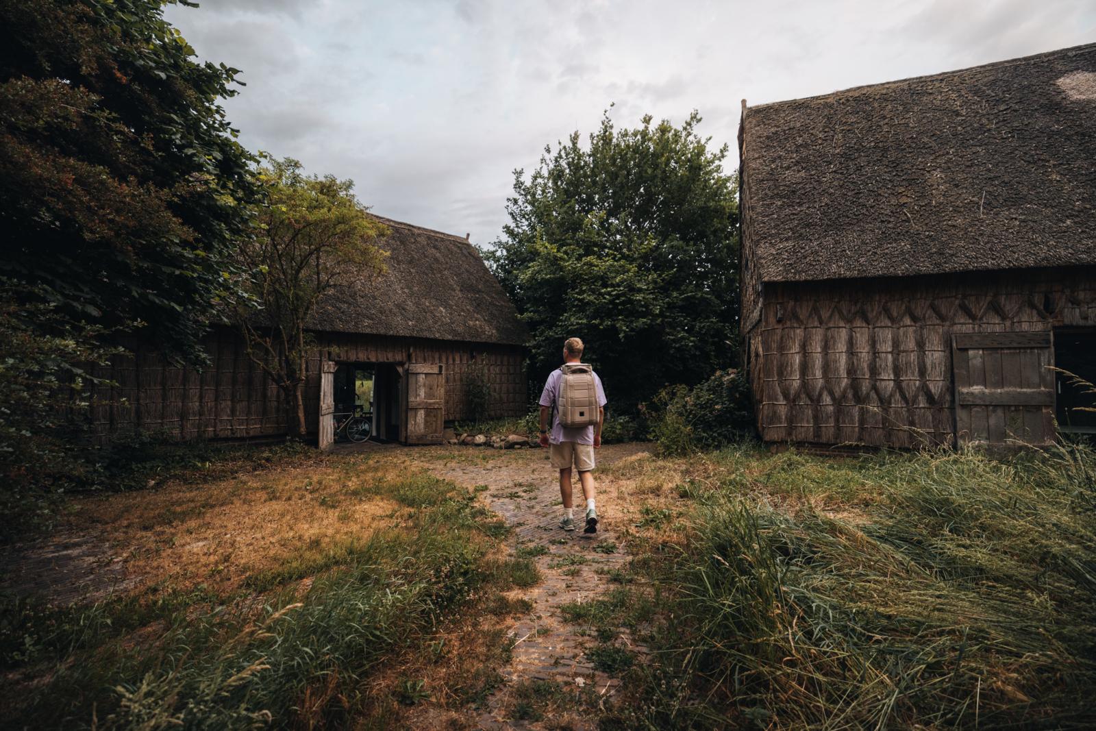 Een wandelaar met rugzak loopt over een onverhard pad naar de historische rietgedekte Wilmsboo boerderij in Drenthe.
