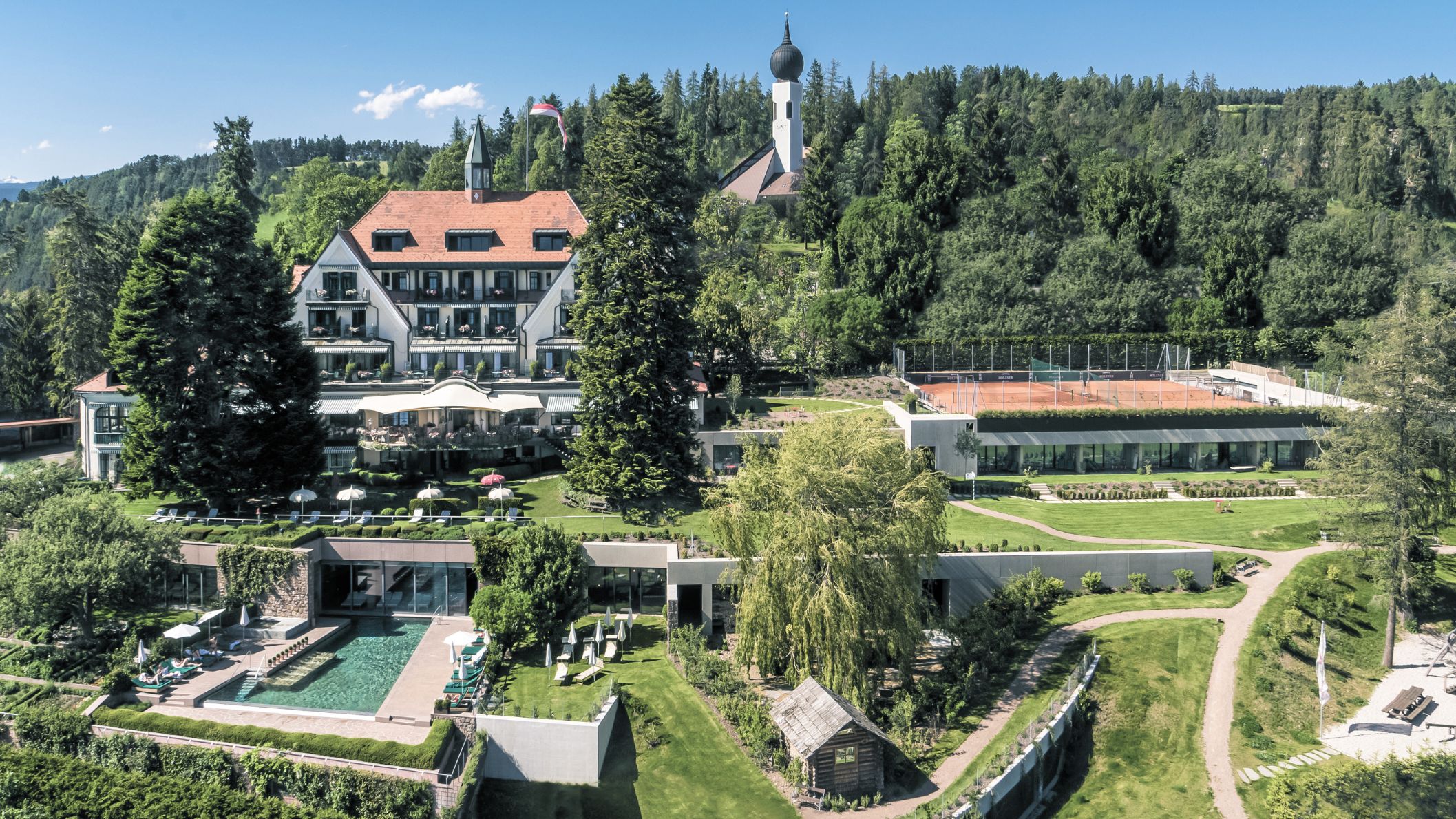 Luchtfoto van een groot hotel met een buitenzwembad, tennisbanen en een kerktoren op de achtergrond.