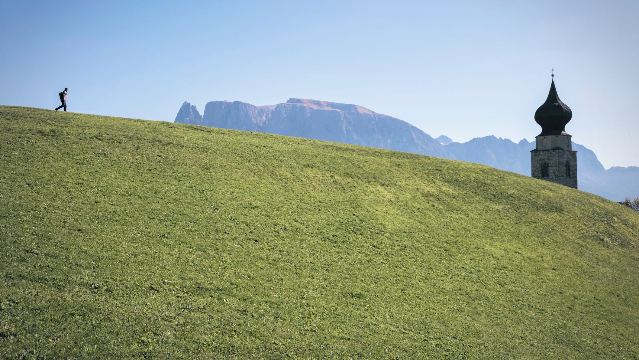 Een eenzame wandelaar loopt over een groene helling voor een berglandschap met een traditionele kerktoren.