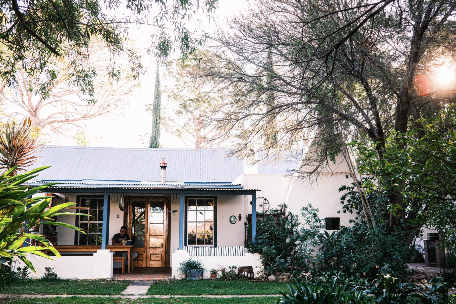 Een charmant, historisch wit huis met veranda in Prince Albert, Zuid-Afrika, omringd door bomen en weelderige tuinbeplanting, met iemand die rustig op de veranda leest.