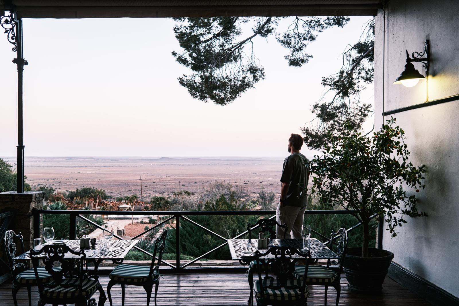 Een persoon staat op een schaduwrijk terras met gedekte tafels en kijkt uit over het uitgestrekte, dorre landschap van de Karoo bij Lemoenfontein Game Lodge in Zuid-Afrika.