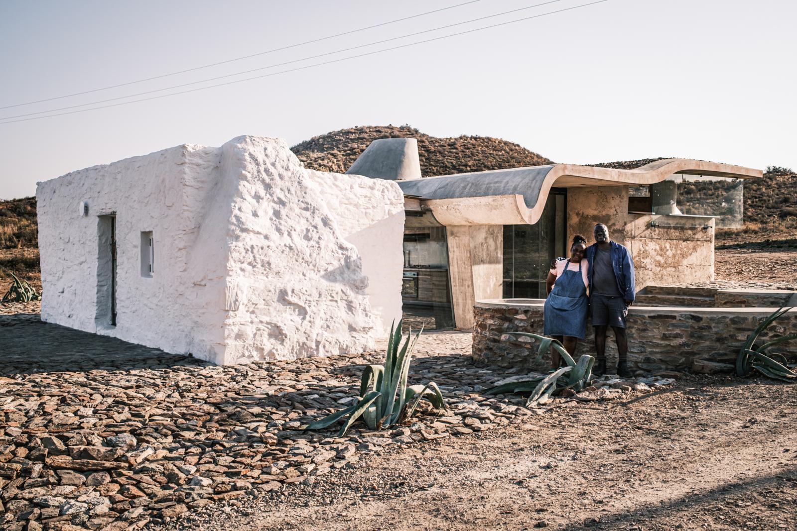 Een koppel poseert voor een uniek gebouw in Buffelsdrift, Zuid-Afrika, waar een traditioneel wit, ruw gestuct huis overgaat in een moderne constructie met een golvend betonnen dak.