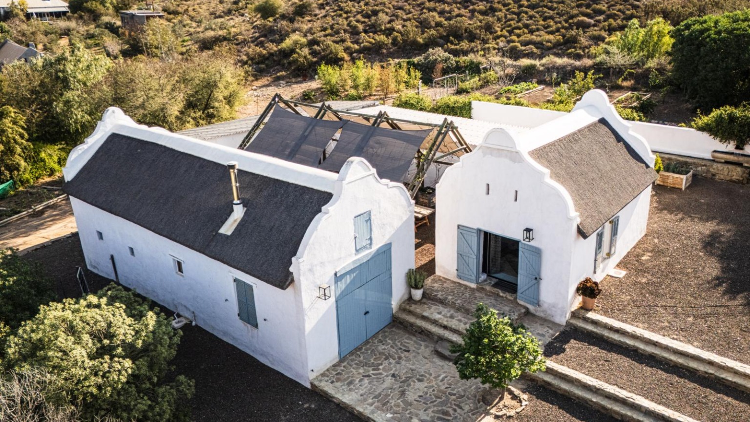 Een bovenaanzicht van een traditionele witte boerderij met rieten dak en blauwe luiken in het landschap van Buffelsdrift in Zuid-Afrika.