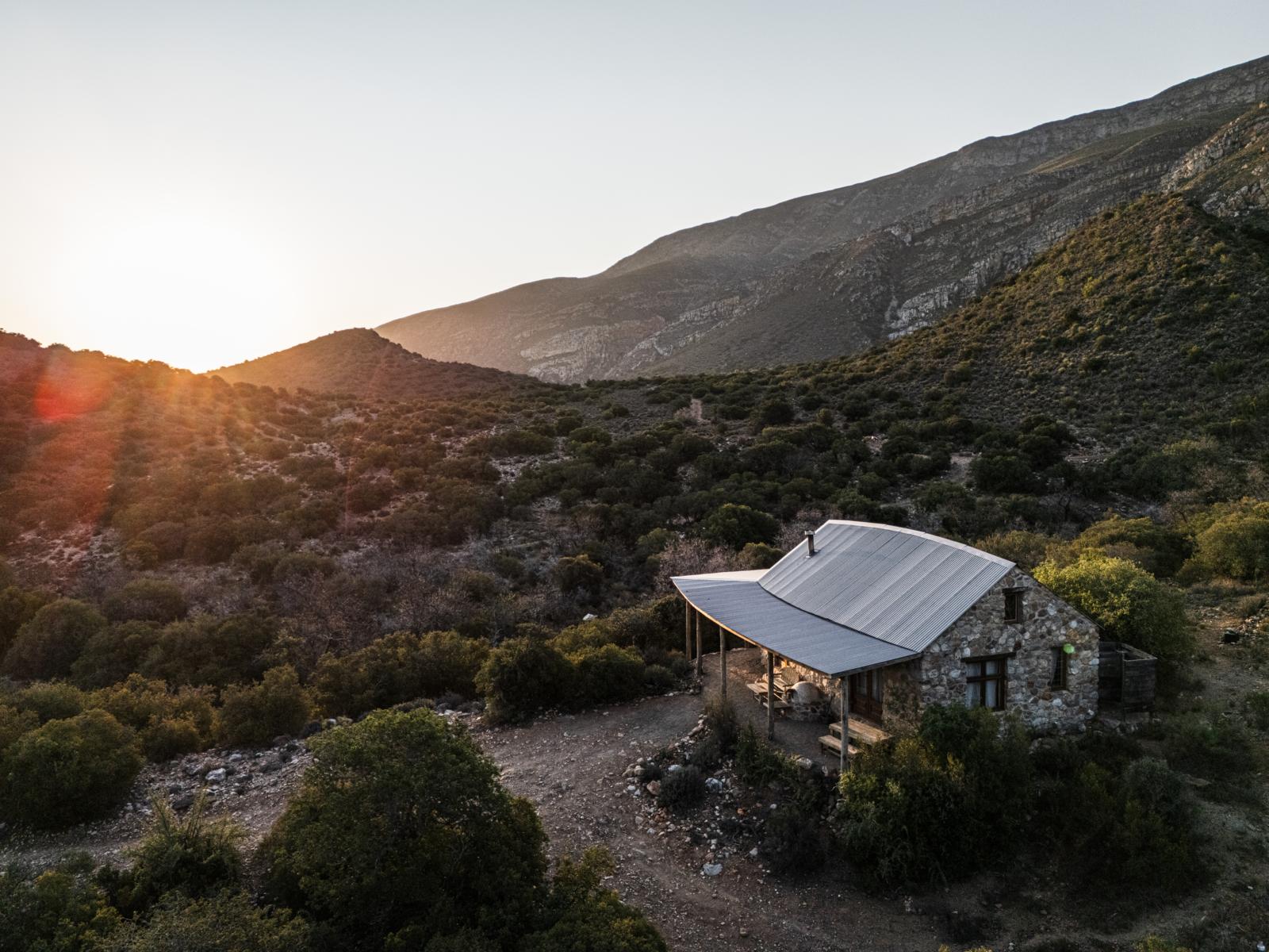 Een stenen lodge met een metalen dak in een uitgestrekt berglandschap van het Touwsberg Private Game and Nature Reserve in Zuid-Afrika, verlicht door de ondergaande zon.