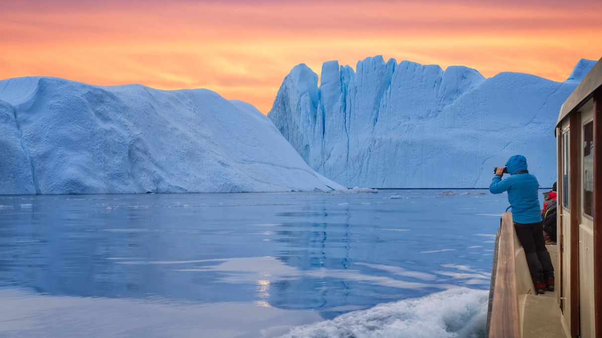 Een toerist in een boot voor een ijsberg, die foto’s maakt bij de zonsondergang tijdens de middernachtzon. Een man, een ontdekkingsreiziger, kijkt vanaf de kust van Ilulissat toe hoe een grote ijsberg in de Arctische zee smelt. Een man verkent in het voorjaar het gebied rond de Ilulissat-ijsfjord in Groenland. Een ontzagwekkende soloreis naar een UNESCO-werelderfgoedlocatie. De Ilulissat-ijsfjord, Disko-baai, Groenland