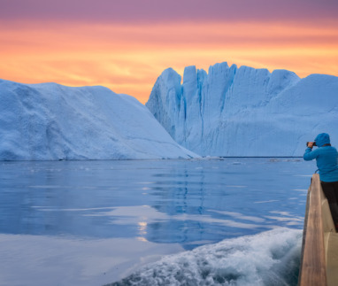 Een toerist in een boot voor een ijsberg, die foto’s maakt bij de zonsondergang tijdens de middernachtzon. Een man, een ontdekkingsreiziger, kijkt vanaf de kust van Ilulissat toe hoe een grote ijsberg in de Arctische zee smelt. Een man verkent in het voorjaar het gebied rond de Ilulissat-ijsfjord in Groenland. Een ontzagwekkende soloreis naar een UNESCO-werelderfgoedlocatie. De Ilulissat-ijsfjord, Disko-baai, Groenland