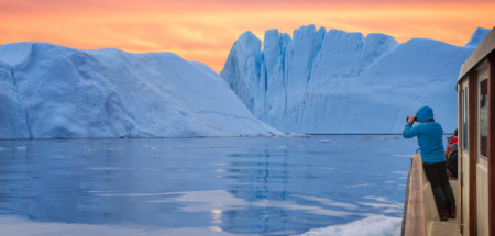 Een toerist in een boot voor een ijsberg, die foto’s maakt bij de zonsondergang tijdens de middernachtzon. Een man, een ontdekkingsreiziger, kijkt vanaf de kust van Ilulissat toe hoe een grote ijsberg in de Arctische zee smelt. Een man verkent in het voorjaar het gebied rond de Ilulissat-ijsfjord in Groenland. Een ontzagwekkende soloreis naar een UNESCO-werelderfgoedlocatie. De Ilulissat-ijsfjord, Disko-baai, Groenland