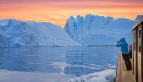 Een toerist in een boot voor een ijsberg, die foto’s maakt bij de zonsondergang tijdens de middernachtzon. Een man, een ontdekkingsreiziger, kijkt vanaf de kust van Ilulissat toe hoe een grote ijsberg in de Arctische zee smelt. Een man verkent in het voorjaar het gebied rond de Ilulissat-ijsfjord in Groenland. Een ontzagwekkende soloreis naar een UNESCO-werelderfgoedlocatie. De Ilulissat-ijsfjord, Disko-baai, Groenland