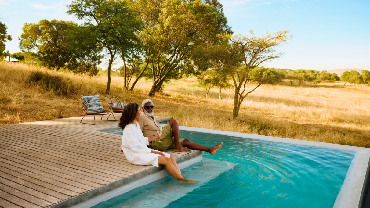 Een volwassen stel geniet van een luxe safarivakantie en laat hun voeten in een zwembad met uitzicht op de savanne hangen