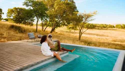Een volwassen stel geniet van een luxe safarivakantie en laat hun voeten in een zwembad met uitzicht op de savanne hangen