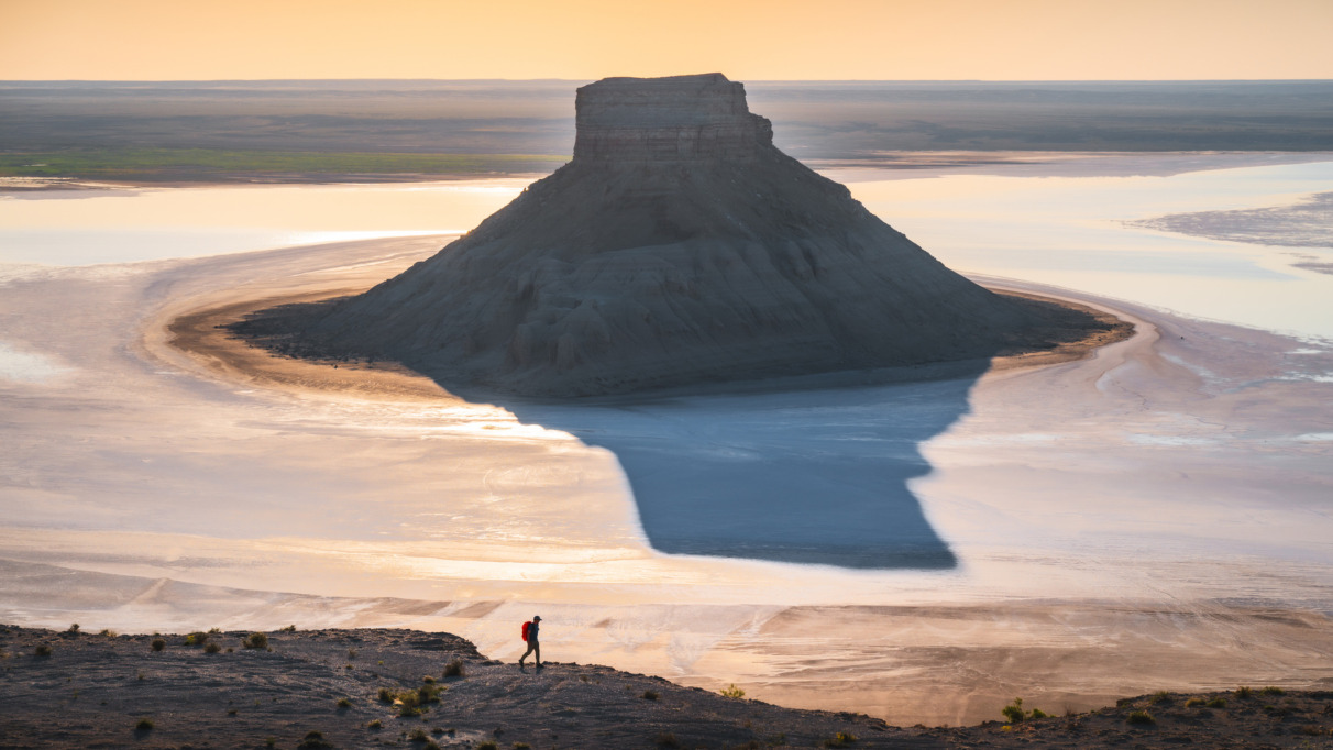 Een kegelvormige rotsheuvel rijst op uit een spiegelglad zoutmeer waarin het gouden licht van de zonsopgang weerspiegelt, terwijl een kleine wandelaar over een bergkam op de voorgrond loopt, in de regio Mangystau, in het westen van Kazachstan.