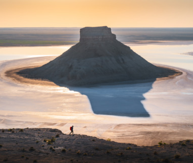 Een kegelvormige rotsheuvel rijst op uit een spiegelglad zoutmeer waarin het gouden licht van de zonsopgang weerspiegelt, terwijl een kleine wandelaar over een bergkam op de voorgrond loopt, in de regio Mangystau, in het westen van Kazachstan.