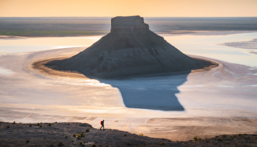 Een kegelvormige rotsheuvel rijst op uit een spiegelglad zoutmeer waarin het gouden licht van de zonsopgang weerspiegelt, terwijl een kleine wandelaar over een bergkam op de voorgrond loopt, in de regio Mangystau, in het westen van Kazachstan.