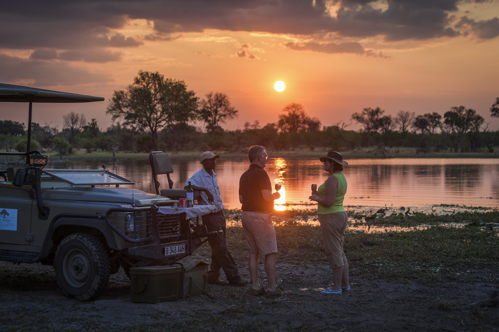 Toeristen genieten van een drankje bij zonsondergang aan de oever van de Khwai-rivier. Okavango-delta. Machaba-kamp. Botswana