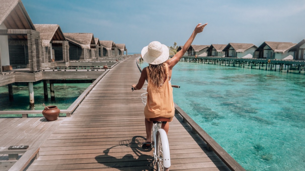 Tropische vakantie: een jonge vrouw op een fiets op een houten steiger op de Malediven, terwijl ze de villa’s boven het water op het eiland bewondert. Een vrouw geniet van een fietstochtje op een steiger boven het water van het koraalrif. Een sprookjesachtige bestemming