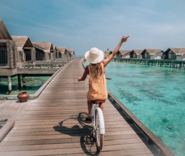 Tropische vakantie: een jonge vrouw op een fiets op een houten steiger op de Malediven, terwijl ze de villa’s boven het water op het eiland bewondert. Een vrouw geniet van een fietstochtje op een steiger boven het water van het koraalrif. Een sprookjesachtige bestemming
