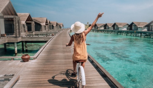 Tropische vakantie: een jonge vrouw op een fiets op een houten steiger op de Malediven, terwijl ze de villa’s boven het water op het eiland bewondert. Een vrouw geniet van een fietstochtje op een steiger boven het water van het koraalrif. Een sprookjesachtige bestemming