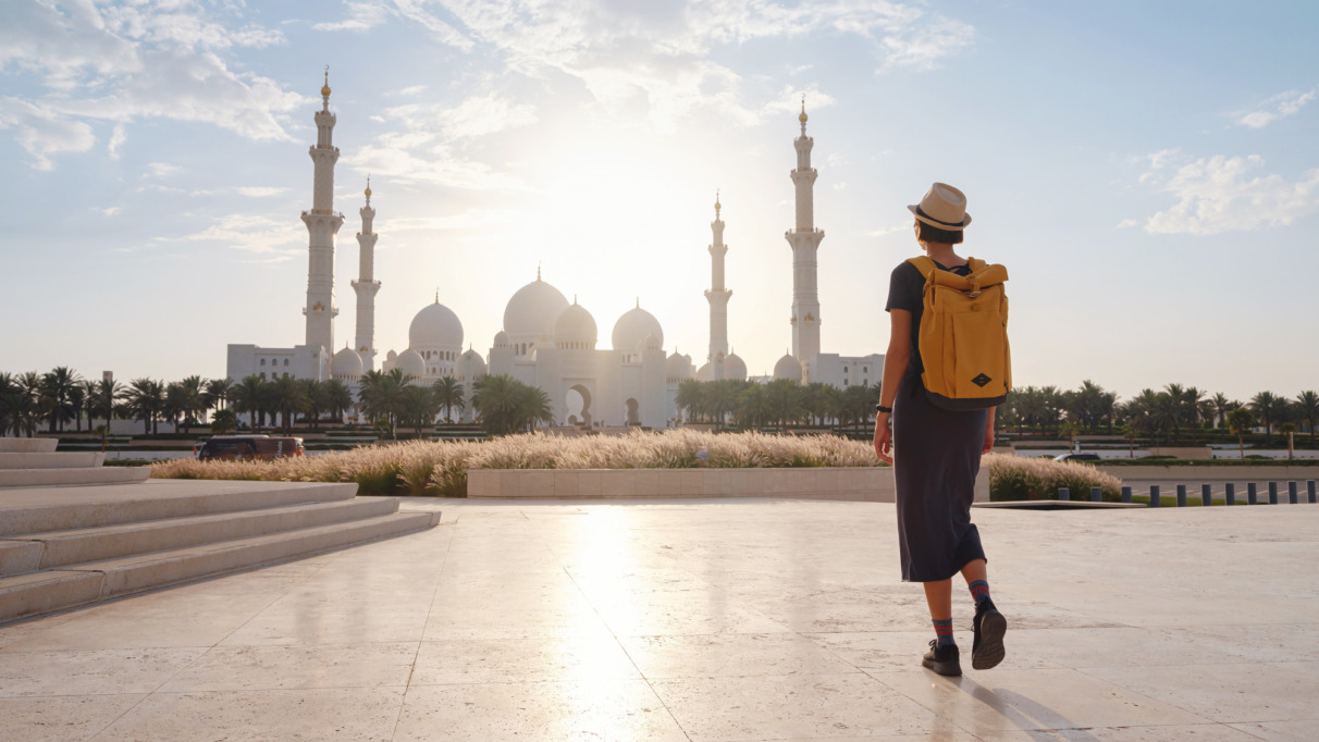 Reis naar de Verenigde Arabische Emiraten: een vrolijke jonge Aziatische reizigster met rugzak en hoed in Wahat Al Karama, ook wel de ‘Oase van Waardigheid’ genoemd, een permanent gedenkteken voor de martelaren, en de Shaikh Zayed Grand Mosque.