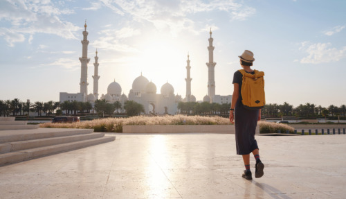 Reis naar de Verenigde Arabische Emiraten: een vrolijke jonge Aziatische reizigster met rugzak en hoed in Wahat Al Karama, ook wel de ‘Oase van Waardigheid’ genoemd, een permanent gedenkteken voor de martelaren, en de Shaikh Zayed Grand Mosque.