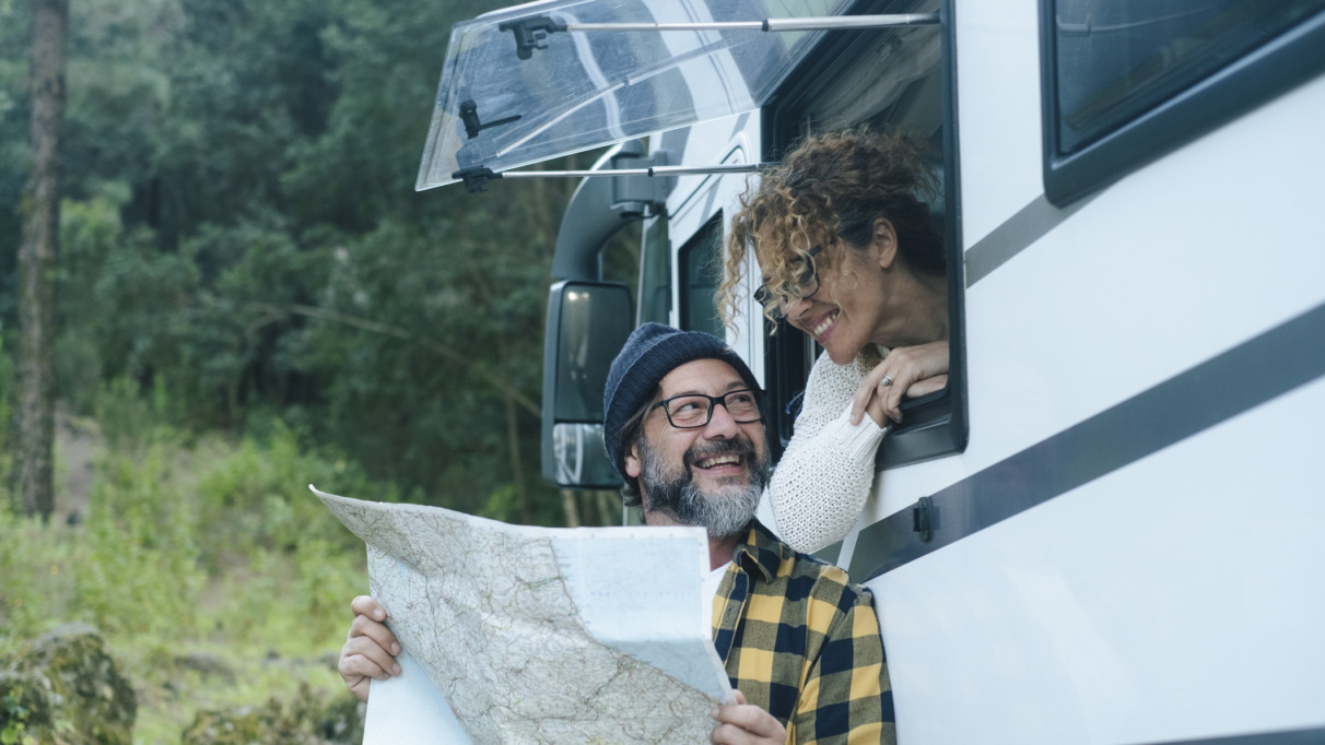 Een gelukkig, volwassen stel geniet van het camperleven: ze reizen, praten en bekijken samen een kaart om hun volgende vakantiebestemming te kiezen. De man staat buiten en de vrouw zit bij het raam. Camper