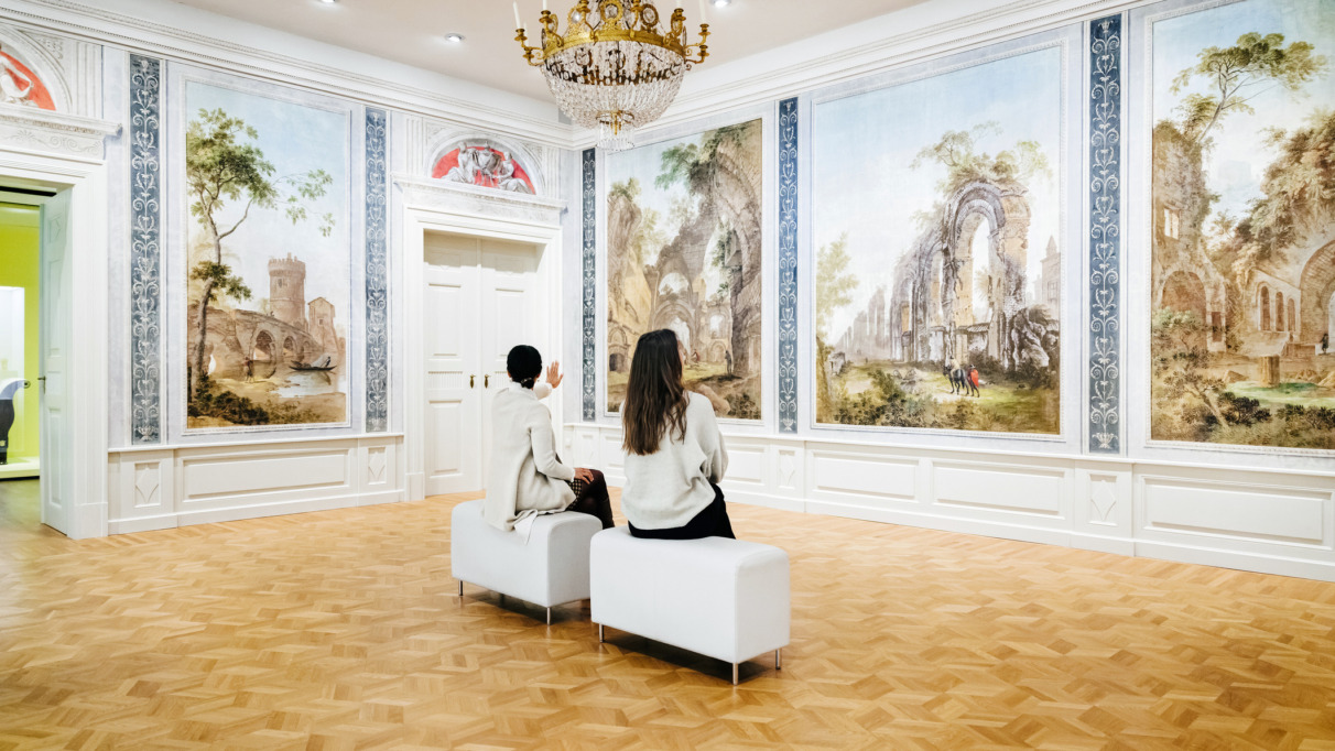 Twee vrouwen zitten op bankjes in een grote galerij en zijn volledig in beslag genomen door het bekijken en bespreken van de klassieke schilderijen die de muren sieren.