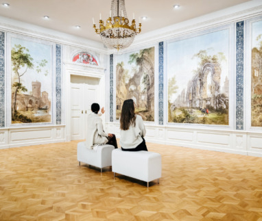 Twee vrouwen zitten op bankjes in een grote galerij en zijn volledig in beslag genomen door het bekijken en bespreken van de klassieke schilderijen die de muren sieren.