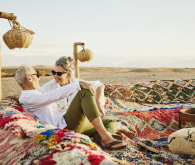 Breedbeeldopname van een glimlachend bejaard echtpaar dat tijdens hun vakantie in Marokko ontspant en geniet van de zonsondergang in een luxe woestijnkamp