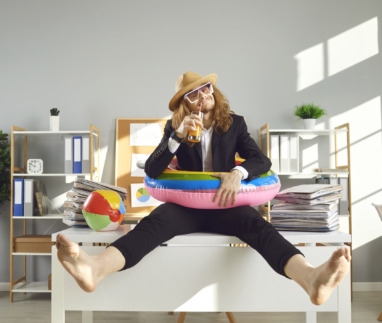 Grappige kantoormedewerker die uitkijkt naar de zomervakantie. Vrolijke medewerker op blote voeten, met een zonnehoed, zonnebril en een zwemring, zittend op het bureau en een cocktail drinkend