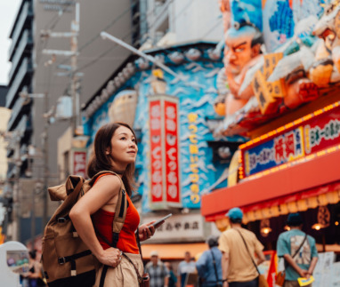 Een jonge Aziatische vrouw die een kaart-app met locatietracking op haar smartphone gebruikt terwijl ze door een straat in Shinsekai, Osaka, Japan, slentert. Ze draagt een rugzak en een rood hemdje. Reizigers die de Japanse cultuur ontdekken.