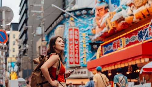 Een jonge Aziatische vrouw die een kaart-app met locatietracking op haar smartphone gebruikt terwijl ze door een straat in Shinsekai, Osaka, Japan, slentert. Ze draagt een rugzak en een rood hemdje. Reizigers die de Japanse cultuur ontdekken.