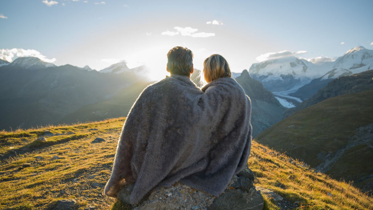 Een stel zit dicht tegen elkaar aan onder een deken en kijkt naar de zonsopgang boven de bergen in Zwitserland