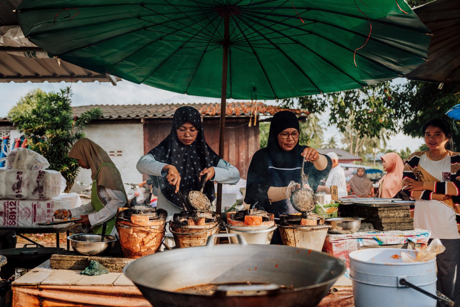 Twee lokale vrouwen in hoofddoek die traditioneel straatvoedsel bereiden op een markt in Nakhon Si Thammarat, Zuid-Thailand, onder een grote groene parasol.